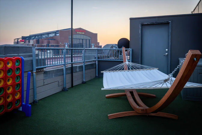 Cozy rooftop patio at sunset with a wooden-frame hammock on artificial turf, oversized Connect Four game, metal railing and views of a nearby brick sports stadium and urban skyline.