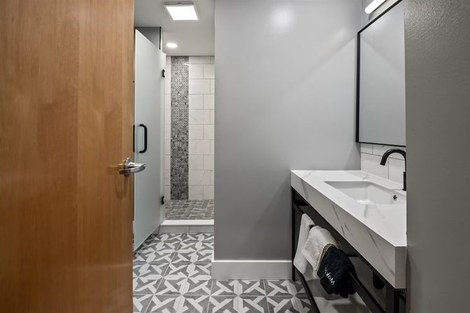 Contemporary gray bathroom interior with geometric patterned floor tiles, frosted glass walk-in shower with mosaic accent strip, white marble rectangular sink on a black metal vanity and matte black faucet