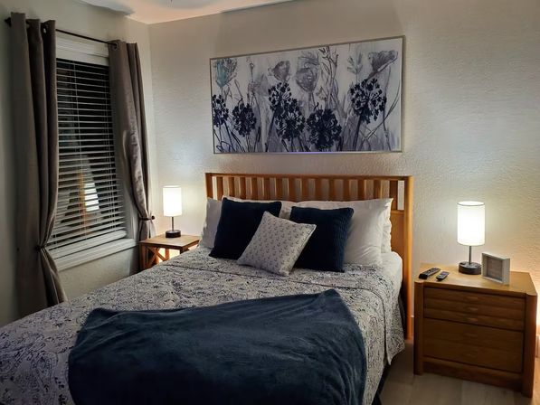 Cozy modern bedroom with a wooden slat bed, patterned duvet, navy throw and pillows, matching wooden nightstands with table lamps, floral wall art above the bed, and a window with blinds and neutral curtains.