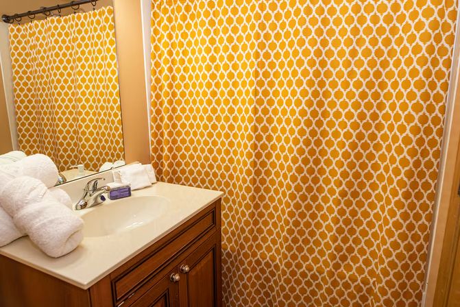 Bright bathroom with yellow quatrefoil-pattern shower curtain, white sink set in a wooden vanity, chrome faucet, bar soap and rolled white towels by a mirror.