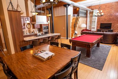 Open-plan urban loft interior with exposed brick wall, hardwood floors, kitchen island with bar stools, wooden dining table, red felt pool table, leather sofa and lofted bedroom.