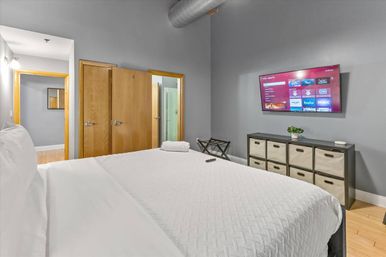 Cozy modern urban loft bedroom with white quilted bedding, wall-mounted smart TV showing streaming apps, gray walls, exposed ducting, wooden doors, fabric-bin dresser and hardwood floor.