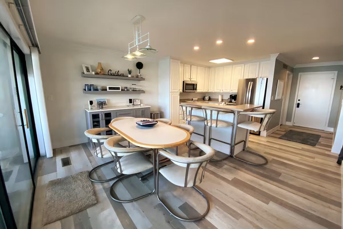 Bright open-plan kitchen and dining area with white cabinets and stainless-steel appliances, a breakfast bar with metal stools, oval dining table surrounded by curved metal chairs, sliding glass patio doors, pendant lights and light wood-look flooring