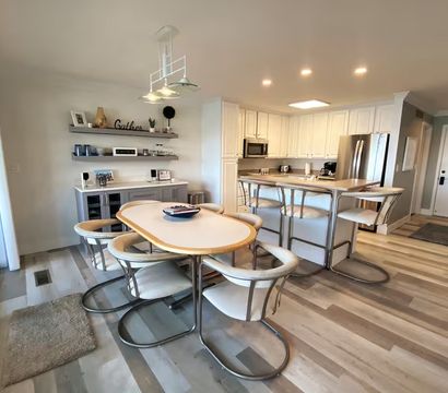 Bright open-plan kitchen and dining area with white cabinets and stainless-steel appliances, a breakfast bar with metal stools, oval dining table surrounded by curved metal chairs, sliding glass patio doors, pendant lights and light wood-look flooring