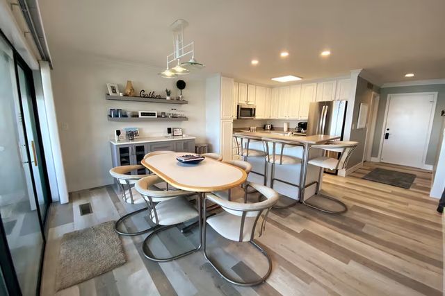 Bright open-plan kitchen and dining area with white cabinets and stainless-steel appliances, a breakfast bar with metal stools, oval dining table surrounded by curved metal chairs, sliding glass patio doors, pendant lights and light wood-look flooring