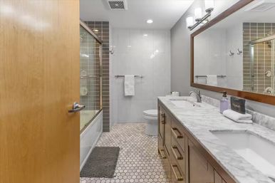Modern residential bathroom interior with marble double vanity and sinks, large framed mirror, glass-enclosed tub with gray subway tile, hexagon mosaic floor, and wooden door slightly ajar.
