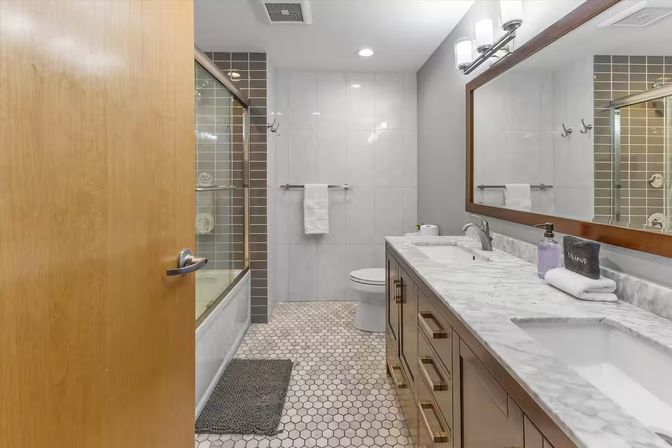 Modern residential bathroom interior with marble double vanity and sinks, large framed mirror, glass-enclosed tub with gray subway tile, hexagon mosaic floor, and wooden door slightly ajar.