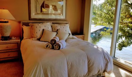 Sunlit lakefront bedroom with plush white bedding, decorative pillows and three rolled towels on the bed, wooden nightstand and floor-to-ceiling window overlooking a dock and leafy tree over calm water.