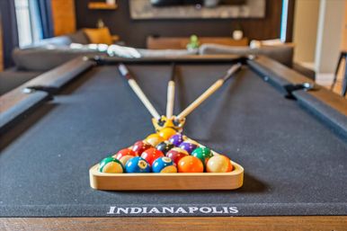 Black-felt pool table in Indianapolis with a racked triangle of colorful billiard balls and three cue sticks crossed behind them in a cozy game room