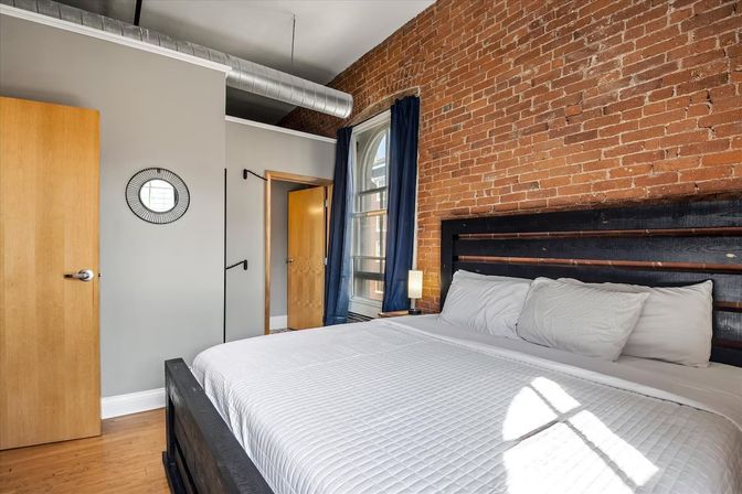 Cozy sunlit urban loft bedroom with exposed brick wall, king bed with white quilt and dark wooden headboard, arched window with navy curtains, hardwood floor and visible industrial ductwork.