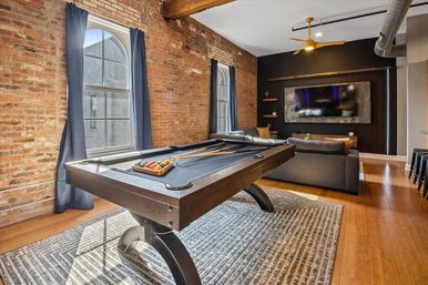 Industrial loft game room with dark pool table and cues, exposed brick walls, arched windows with blue curtains, gray sectional, wall-mounted TV, wood floors, modern ceiling fan