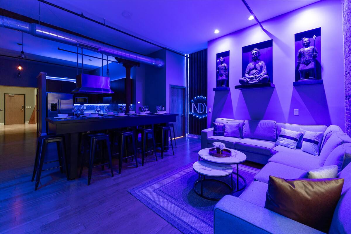 Open-plan urban loft living room and kitchen with industrial ductwork, long black bar and stools, stainless appliances, purple-blue LED lighting, cozy sectional sofa, nested marble coffee tables, and three illuminated Buddha statues in wall niches.