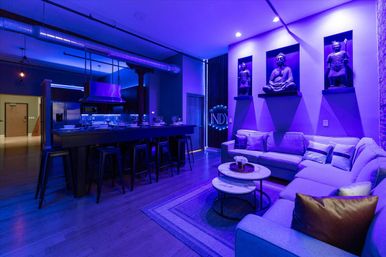 Open-plan urban loft living room and kitchen with industrial ductwork, long black bar and stools, stainless appliances, purple-blue LED lighting, cozy sectional sofa, nested marble coffee tables, and three illuminated Buddha statues in wall niches.