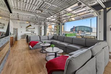 Cozy modern rooftop lounge with a large gray sectional, wood floors, round coffee tables and open garage-style doors revealing an outdoor terrace and downtown arena view.