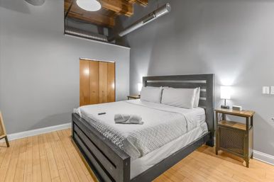 Cozy modern urban loft bedroom with exposed ductwork and wooden ceiling beams, king bed dressed in white linens, bedside tables with lamps, gray walls and light hardwood floors.