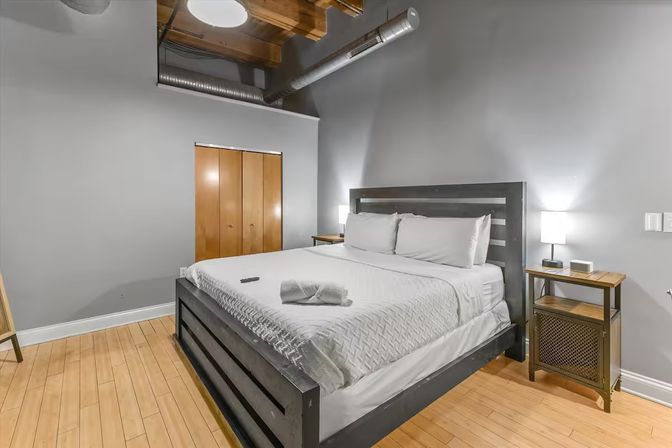 Cozy modern urban loft bedroom with exposed ductwork and wooden ceiling beams, king bed dressed in white linens, bedside tables with lamps, gray walls and light hardwood floors.