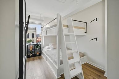Bright modern urban apartment bedroom with white wooden bunk bed and ladder, light oak floors, city-building view from the window, black cube shelf with plants and colorful boxes, minimalist white walls and wall hooks.