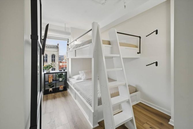 Bright modern urban apartment bedroom with white wooden bunk bed and ladder, light oak floors, city-building view from the window, black cube shelf with plants and colorful boxes, minimalist white walls and wall hooks.