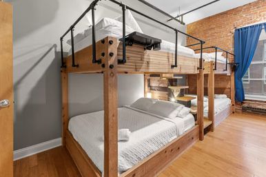 Cozy industrial-loft bunk room with solid wood double-decker beds, crisp white linens, exposed red brick wall, large window with blue curtain and warm hardwood floors