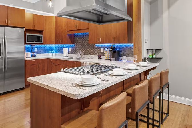 Modern open-plan kitchen with speckled quartz island and gas cooktop, four leather bar stools, stainless-steel refrigerator, warm wood cabinets and blue LED tile backsplash.