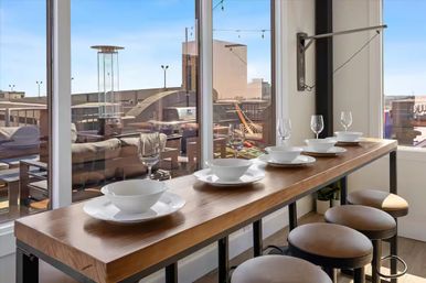 Sleek wooden counter set with bowls and wine glasses, bar stools by floor-to-ceiling windows overlooking a sunny urban rooftop terrace and downtown skyline.