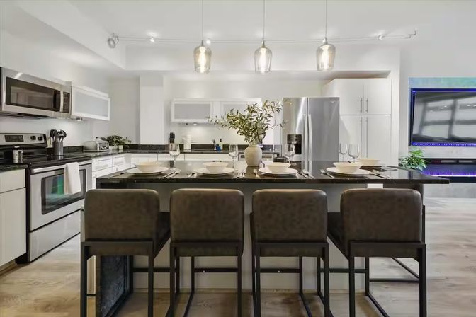 Stylish urban loft kitchen with a black granite island set for four, leather bar stools, stainless-steel appliances, white cabinets, glass pendant lights, and a vase centerpiece
