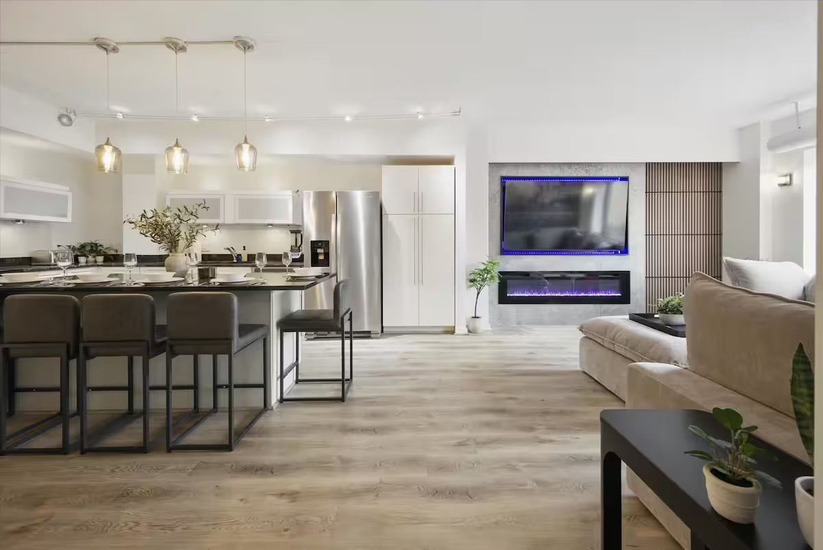 Bright modern open-concept apartment interior with kitchen island and bar stools, stainless steel fridge, pendant lights, light wood floors, LED-lit wall TV and electric fireplace.