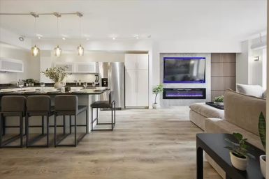 Bright modern open-concept apartment interior with kitchen island and bar stools, stainless steel fridge, pendant lights, light wood floors, LED-lit wall TV and electric fireplace.