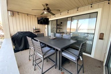 Cozy covered waterfront balcony patio with a dark metal high-top dining table and six cushioned chairs, ceiling fan, string lights, covered grill and sliding glass doors reflecting a river view.