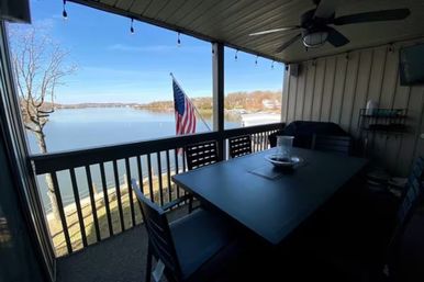 Cozy lakefront covered balcony with patio dining table and chairs, American flag on the railing overlooking a calm lake and tree-lined shoreline under a clear blue sky.