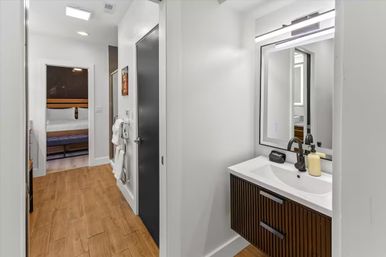 Bright modern hallway with wood-look flooring leading to a bedroom, compact bathroom on the right featuring a dark wood slatted vanity, white sink with black faucet, rectangular mirror and wall-mounted towel rack.