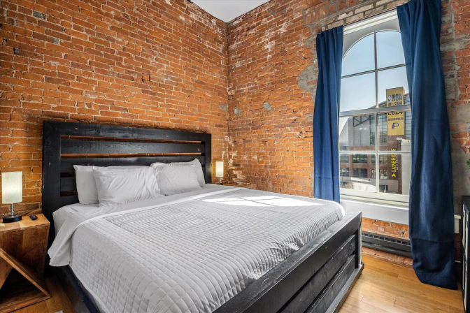 Bright urban loft bedroom with exposed brick walls, large arched window and navy curtains, king bed on a dark wooden frame and hardwood floors.