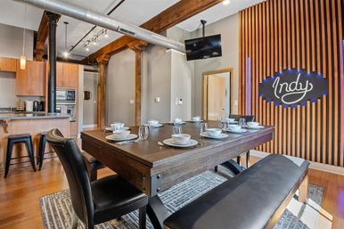 Stylish industrial loft dining room set for eight — heavy reclaimed wood table with leather chairs and bench, open kitchen, exposed wooden beams and metal ductwork, and a wood-slatted accent wall with an illuminated 'Indy' sign.