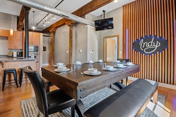 Stylish industrial loft dining room set for eight — heavy reclaimed wood table with leather chairs and bench, open kitchen, exposed wooden beams and metal ductwork, and a wood-slatted accent wall with an illuminated 'Indy' sign.