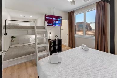 Bright modern downtown vacation rental bedroom with white built-in bunk beds, queen bed topped with folded towels, wall-mounted TV, hardwood floors and large window with city view.