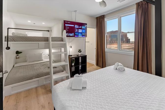 Bright modern downtown vacation rental bedroom with white built-in bunk beds, queen bed topped with folded towels, wall-mounted TV, hardwood floors and large window with city view.