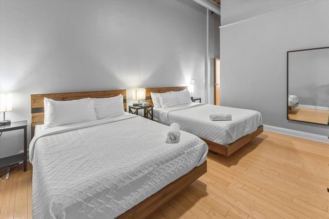 Bright modern hotel room with two large beds topped with white bedding and rolled towels, wooden headboards, bedside lamps and nightstands, gray walls, hardwood floor and full-length mirror.