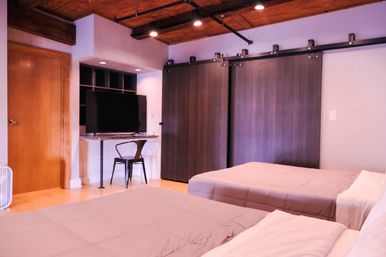 Cozy modern loft-style bedroom with exposed wood ceiling and track lighting, two beds, small desk with TV and metal chair, and dark sliding barn doors.