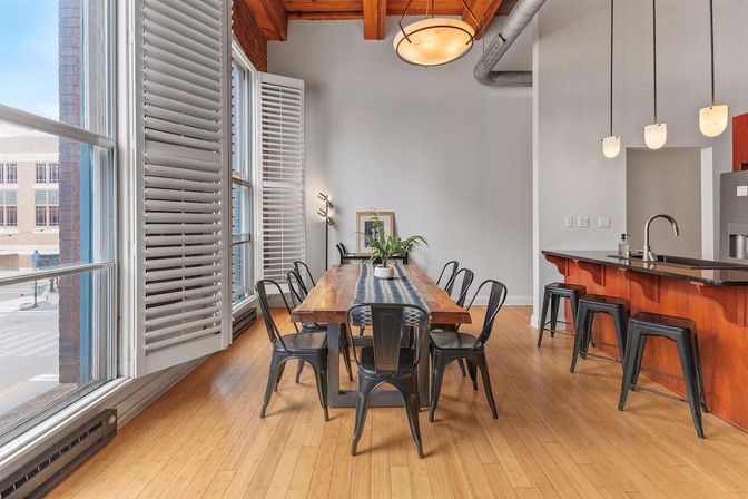 Sunlit urban loft dining area with a long wooden farmhouse table topped with a blue runner and potted plant, black metal chairs, floor-to-ceiling shuttered windows, exposed wood beams, hardwood floors, and a kitchen island with pendant lights and bar stools.