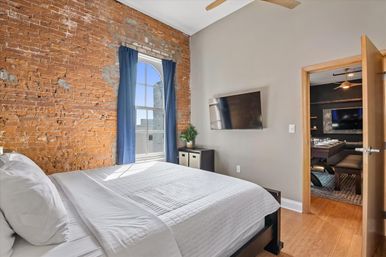 Cozy sunlit urban loft bedroom with exposed brick wall, arched window framed by blue curtains, white quilted bed, wall-mounted TV, and warm wood floors.