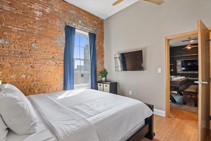 Cozy sunlit urban loft bedroom with exposed brick wall, arched window framed by blue curtains, white quilted bed, wall-mounted TV, and warm wood floors.