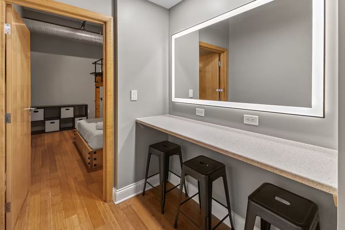Loft-style dressing area with backlit vanity mirror, long light countertop, three black metal stools, hardwood floors and an open wooden door revealing a simple bedroom with low bed and cube storage.