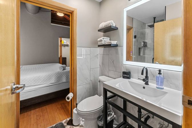 Contemporary bathroom with marble tile, LED-lit mirror, white sink and black faucet, toilet, floating shelves with folded towels, and open door to a bedroom with hardwood floor, bright tidy layout