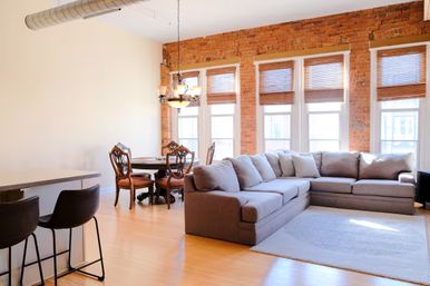 Sunlit open-plan loft living room with exposed brick wall and tall windows, gray L-shaped sectional, wooden dining set and bar stools on light hardwood floors — modern-industrial cozy vibe.