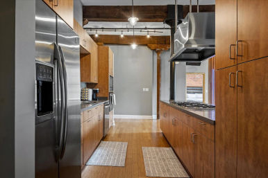 Modern industrial galley kitchen — stainless refrigerator and hood, warm wood cabinets, gas cooktop, exposed wooden beams and hardwood floors.