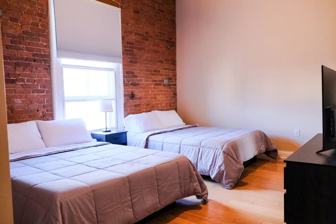 Cozy urban loft bedroom with exposed red brick wall, hardwood floors, two neatly made beds, bedside table and lamp, and a TV on a dresser