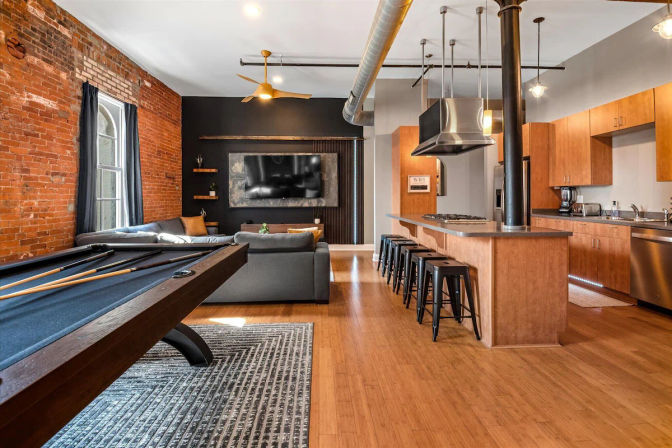 Downtown open-plan loft apartment with exposed brick wall, hardwood floors, pool table, gray sectional facing a wall-mounted TV, industrial ductwork and ceiling fan, and a modern kitchen island with black bar stools and stainless steel appliances.