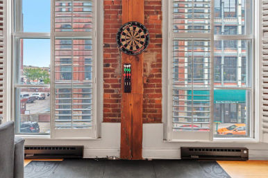 Loft apartment interior with exposed brick and wooden beam holding a dartboard, flanked by tall white shuttered windows overlooking a city street with parked cars and brick storefronts