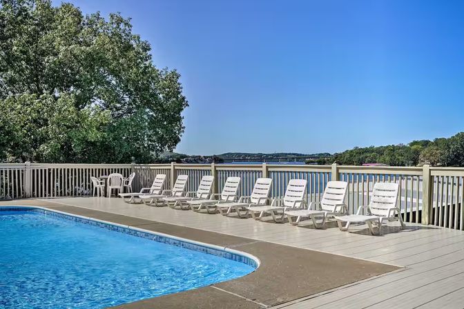 Lakeside pool deck with a row of white lounge chairs along a wooden railing, calm water and trees under a clear blue sky