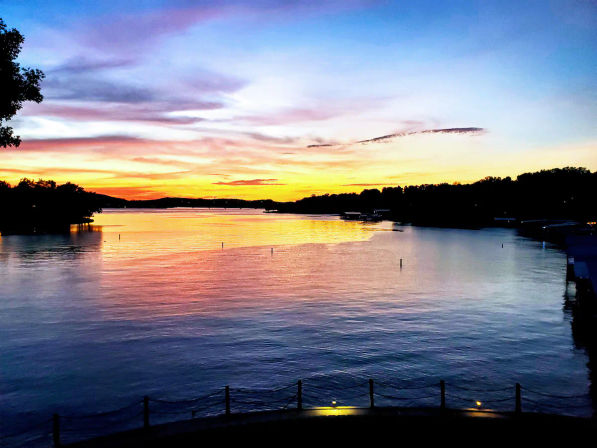 Vibrant sunset over a calm lake, orange, pink and blue sky reflected on the water with silhouetted tree-lined shoreline and small docks in the distance.